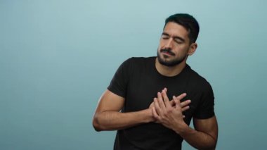 Mavi arka planda göğüs ağrısı çeken genç bir adam, ellerinin göğsünü tutarak rahatsızlığını ve endişesini iletiyor..