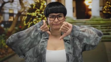 Kadın açık havada başparmakları aşağıda, festival ışıkları ve merdivenlerinin önünde dikilirken, şehir ortamında memnuniyetsizliğini dile getiriyor..