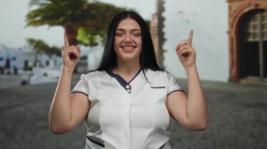 Açık havada gülümseyen bir kadın işaret parmaklarıyla şehrin arka planına doğru bakıyor..