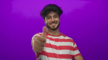 Sakallı, çizgili tişörtlü ve gözlüklü genç adam canlı mor arka planda parmaklarıyla güvenle gülümsüyor..