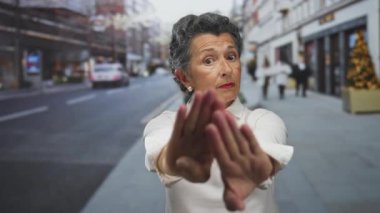 Gri saçlı yaşlı bir kadın, şehir sokaklarında beyaz bir gömlek giyerek bir dur hareketi yapıyor. Şehir dışındaki bir çevrede ihtiyatı ve vurguyu gösteriyor..