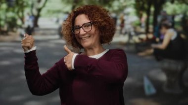 Kıvırcık esmer saçlı ve gözlüklü İspanyol bir kadın. Neşeli bir parkta güneşli bir günün tadını çıkarırken neşe içinde parmaklarını havaya doğrultuyor..