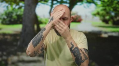 Sakallı dövmeli kel bir adam, güneşli bir günde parkta şakacı bir şekilde yüzünü gizliyor ve ortaya çıkarıyor. Kollarında ve ellerinde karmaşık dövmeler gösteriyor..