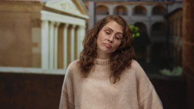 Eski bir üniversitede gözlüklü ve kıvırcık saçlı bir süveter giyen bir kadın klasik mimariyle çevrili düşünceli bir ifade sergiliyor..