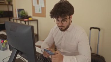 Sakallı ve gözlüklü genç adam bilgisayar ve bavulla donatılmış modern bir ofis yerinde çalışırken akıllı telefonu araştırıyor..