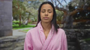 Pembe bornozlu bir kadın yeşil park çevresiyle çevrili düşünceyi gösteren jestler yapıyor. Rahatlamış ama derin düşüncelere dalmış bir anı yakalıyor..