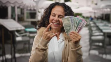 Orta yaşlı bir kadın Hindistan 'da bir restoranın terasında gülümsüyor ve elinde Hint rupisi banknotları tutarken parmaklarıyla taş gibi bir jest yapıyor..