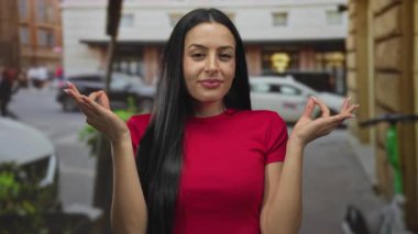 Kırmızı tişörtlü genç İspanyol kadın, kalabalık bir şehir caddesinde arabalarla ve şehir mimarisiyle çevrili, şehir kaosunun ortasında huzur içinde meditasyon yapıyor..