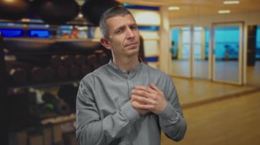 Spor salonundaki adam dingin ifadeyle farkındalık yapıyor, gri gömlek giyiyor, modern fitness ekipmanlarıyla çevrili, sakinliği ve iç mekan gözlemciliğini yansıtıyor..