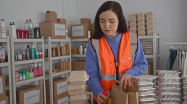Turuncu yelekli bir kadın bağış merkezinde yiyecek paketliyor, raflar erzakla dolu, arka planda organize eşyalar bulunan kapalı alanlarda sosyal yardıma odaklanıyor..