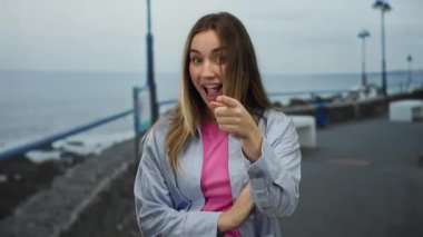 Sarışın kadın deniz kenarındaki bir gezinti alanında komik jestler yapıyor, arka planda okyanus olan eğlenceli bir anı yakalıyor..