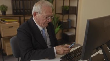 Akıllı telefon kullanan kıdemli bir adam modern ofiste masasında oturuyor ve başarılı haberlerin heyecanını ve neşesini gösteriyor.