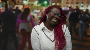 Beyaz süveter içinde gülümseyen kadın canlı şehir caddesinde kalp şekli yaratıyor. Renkli gece ışıkları şehir çeşitliliğini ve neşesini yansıtıyor..