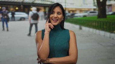 Kalabalık bir caddede gülümseyen genç bir kadın, yeşil bluzlu, arka planda yayalar ve arabalarla kendinden emin ve dostane bir tavır sergiliyor..