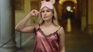 Müzede çılgın bir jest yapan kadın gece elbisesi ve uyku maskesi takıyor, sarı saç, loş ışıklandırma, oyuncu bir ifade ve eşsiz bir atmosferi olan sanat galerisi ayarları..