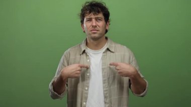 Yüzünde şüphe uyandıran ifadeyle İspanyol bir adam kendine canlı yeşil bir arkaplan işaret ediyor. Görüntüdeki şaşkınlık ve kendini sorgulama duygusunu aktarıyor..