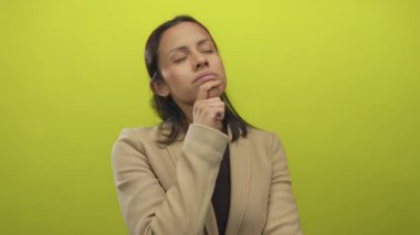 Düşünceli bir kadın, bej ceketli, el pençe divan duran parlak sarı arka planda derin bir tefekkür ve izole bir ortamda iç gözlem öneriyor..