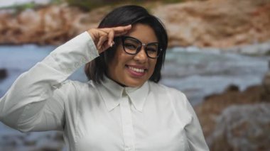 Deniz kenarına gülümseyen kadın selam veriyor. Arkasında dalgalar ve kayalar olan gözlük takıyor.