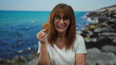 Deniz kenarında gülümseyen İspanyol bir kadın elinde manzaralı bir sahil manzarası ve canlı mavi bir deniz olan bir kurabiye tutuyor..