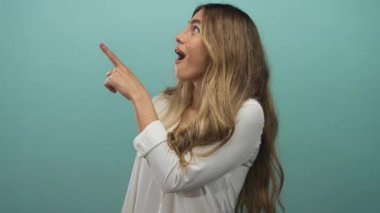 Genç sarışın kadın stüdyo ortamında yukarı doğru işaret ediyor, açık ağızlı ve geniş gözlü çerçeveden dışarı bakıyor; sürpriz merak şaşkınlık.