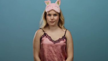 Pembe gecelik ve tavşan uyku maskeli genç sarışın kadın mavi arka planda dikilirken hareketli, neşeli ve dikkatli bir ifade sergiliyor..