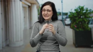 Kadın sokakta duygularını ifade ediyor, elleri kapalı ve gözleri kapalı gri bir kazak giyiyor, esmer saç çerçeveli yüz, yeşillik ve arka planda sütunlar..