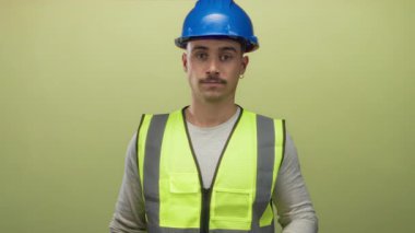 Şapkalı ve yansıtıcı yelekli İspanyol genç adam sarı arka plana karşı yemin ediyor. İnşaattaki bağlılığı ve dürüstlüğü sembolize ediyor..