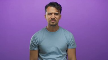 Latin asıllı genç adam orta parmaklarını kaldırarak saldırgan bir jest yapıyor, çarpıcı bir mor arka plana karşı duruyor, meydan okuma ve tavır sergiliyor..