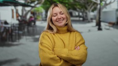Sarı süveter giyen çapraz kollu genç bir kadın şehir terasında gülümsüyor, ağaçlar ve açık hava kafe masaları ile çevrili, kentsel titreşimleri somutlaştıran.