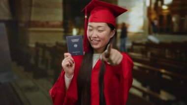 Kırmızı mezuniyet elbisesi giymiş bir kadın kilisede Alman pasaportu taşıyor. Güvenle gülümsüyor ve ileriye bakıyor. Çok kültürlü bir başarıyı ve kimliği temsil ediyor..
