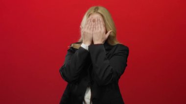 Parlak kırmızı bir arkaplan önünde peekaboo oynayan bir kadın, siyah bir takım elbiseyle karışık sarı saçlar, beklenmedik bir profesyonel ortamda oyunculuk ve entrika çağrıştırıyor..