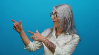 Gri saçlı, dövmeli, gülümseyen ve parlak mavi bir duvara bakan, gözlüklü, beyaz gömlekli, kendine güveni ve tarzı olan son sınıf öğrencisi bir kadın..