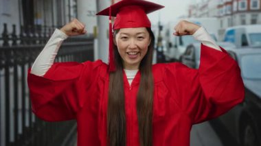 Kırmızı mezuniyet elbisesi giymiş bir kadın kenar mahalle ortamında gururla kaslarını esnetiyor, başarıyı ve başarıyı simgeliyor..