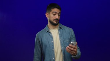 İspanyol asıllı genç adam, özel bir kot gömleği içinde izole edilmiş mavi bir arkaplana karşı akıllı telefon tutarak baş parmak hareketi yapıyor..