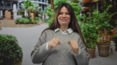 Genç İspanyol kadın yeşil kazaklarla çevrili şehirde neşeli bir ağlama hareketi yapıyor. Canlı bir sokakta eğleniyor ve duygularını canlı bir şekilde ifade ediyor..