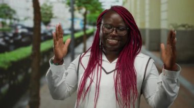 Pembe örgülü bir kadın, şehir caddesinde jest yapıyor, açık havada günlük kıyafetlerle neşe ifade ediyor, enerjik ve arkadaş canlısı ifadesiyle kişiliğini sergiliyor..