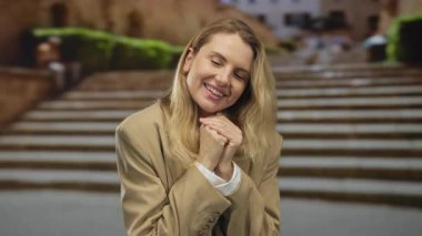 Şehir ortamındaki genç sarışın kadın dışarıda merdivenlerin yanında gülümserken elleriyle kalp hareketi yapıyor..