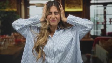 Çizgili gömlekli genç bir kadın, ahşap masanın yanındaki rahat kafenin içinde elleri kulaklarını kapatıyor. Stres baş ağrısı..