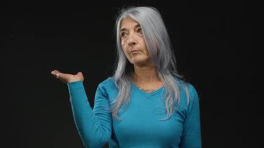 Gri saçlı ve mavi kazaklı bir kadın iki elini kaldırarak derin düşüncelere dalmış ve kafa karışıklığı olduğunu öne sürüyor..