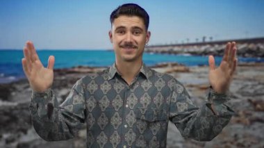 Genç İspanyol adam açık mavi bir deniz ve gökyüzü ile güneşli bir sahil tarafında, açık bir el hareketi yapıyor, desenli bir gömlek giyiyor, neşeli ve rahat bir açık hava atmosferi taşıyor..