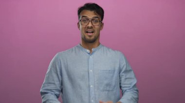 Gözlüklü ve mavi gömlekli genç İspanyol adam, yüz ifadeleri ve jestler kullanarak pembe arka plan üzerinde şaşkınlık ve heyecan ifade ediyor..