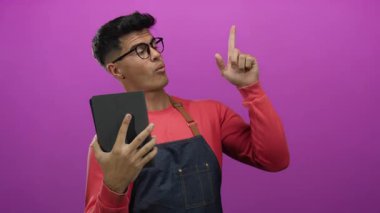 Gözlüklü genç adam elinde tablet, parlak pembe arka plan önlüğü ile derin düşünce ve dikkat çekmeyen bir şeyi işaret ediyor..