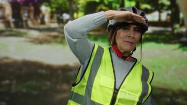 Miğfer ve yansıtıcı yelek giyen kıdemli İspanyol kadın parktaki mesafeye bakıyor. Etrafını çevreleyen ağaçlar ve yeşillikler, dış güvenlik ve yaşlı aktiviteleri somutlaştırıyor..