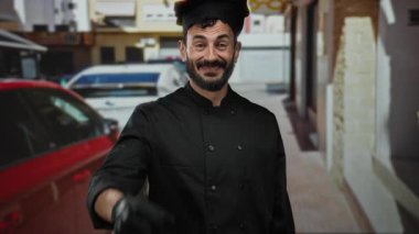 Sakallı İspanyol şef gülümsüyor ve açık havada güneşli bir sokakta siyah üniformalı jest yapıyor..