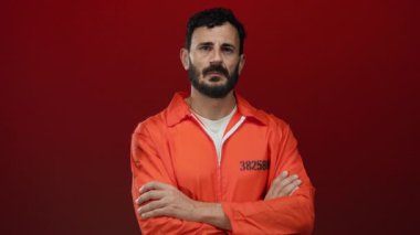 Turuncu hapishane tulumu giyen sakallı İspanyol bir adam koyu kırmızı bir arka plana karşı duruyor ve ciddi bir tavır sergiliyor..