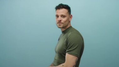 Dövmeleri ve çapraz kolları olan genç adam mavi bir arka plana karşı duruyor, yeşil bir gömlek giyiyor, modern saç stiliyle gülümsüyor, şık ve çekici bir ifade sergiliyor..