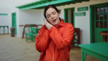 Turuncu tulumlu kadın güneşli terasta poz veriyor, gözleri dingin bir ifadeyle kapalı, yeşil ahşap mobilyalarla çevrili, açık hava restoranında..