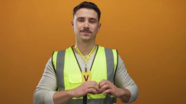 Yansıtıcı yelek giyen genç İspanyol adam turuncu arka plana karşı kalp hareketi yapıyor. Gönüllü desteği ve toplumsal müdahaleyi simgeliyor.