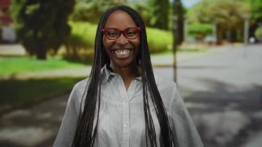 Uzun saçlı bir kadın, şehir parkında neşeli bir şekilde gülümsüyor, güneş ışığı altındaki bir sokakta güvenle duruyor, gözlük ve çizgili bir gömlek giyiyor, mutluluk ve zarafet saçıyor..