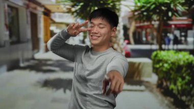 Açık şehir sokak ortamında gülümseyen genç Çinli bir adam hareketli bir şehir ortamında canlı bir yeşilliğin altında canlı bir ifade sergiliyor..